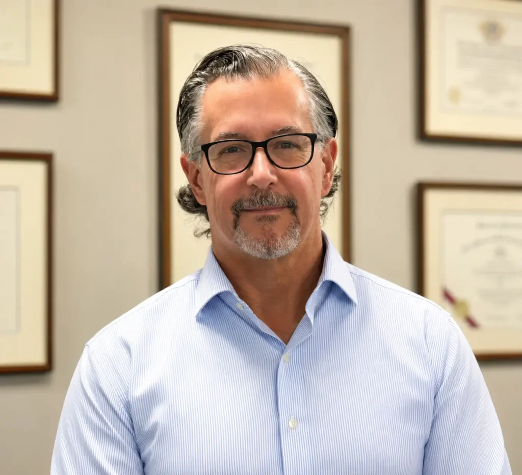 Professional man with glasses smiling in front of framed certificates.